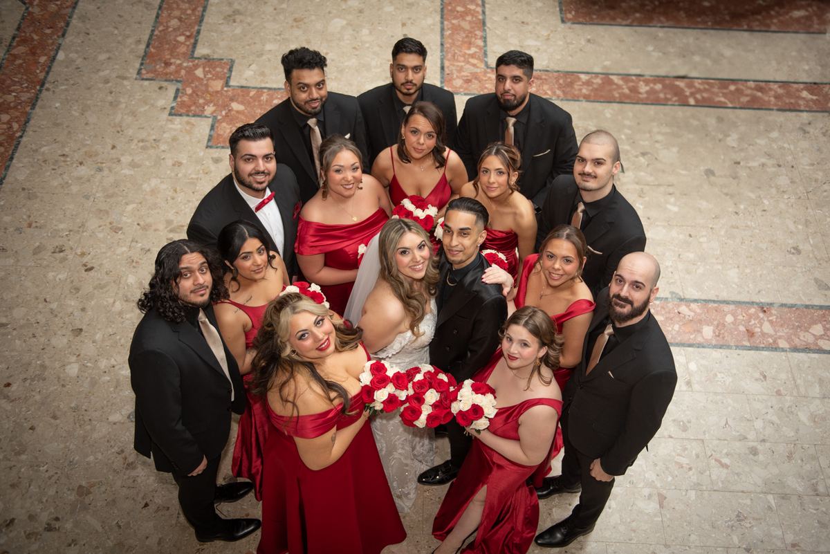 Wedding party in formal attire with bride and groom at center, surrounded by joyful bridesmaids in red satin dresses.