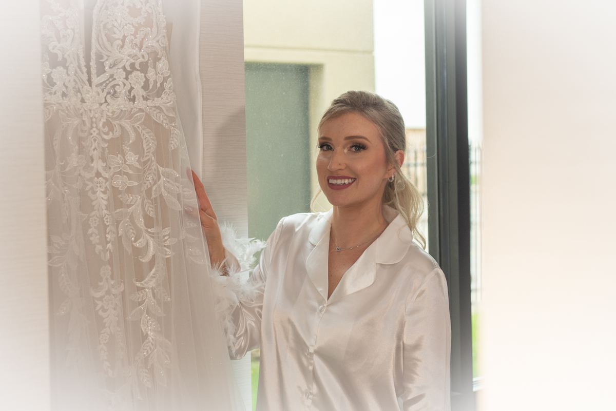 A bride in a satin robe touches her wedding gown hanging beside her in a softly lit room, illuminated by natural light.
