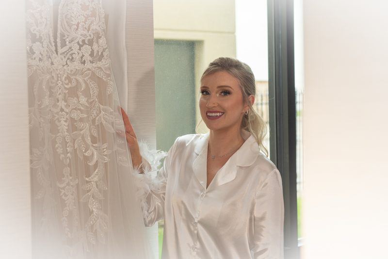 A bride in a satin robe touches her wedding gown hanging beside her in a softly lit room, illuminated by natural light.
