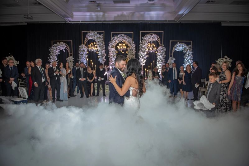 Bride and groom dance on foggy stage surrounded by smiling guests in formal wear, illuminated by chandeliers.