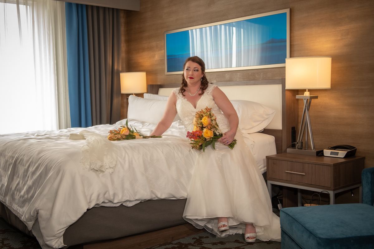 A bride in a white gown sits on the edge of a hotel bed, holding an arrangement of orange and yellow flowers.