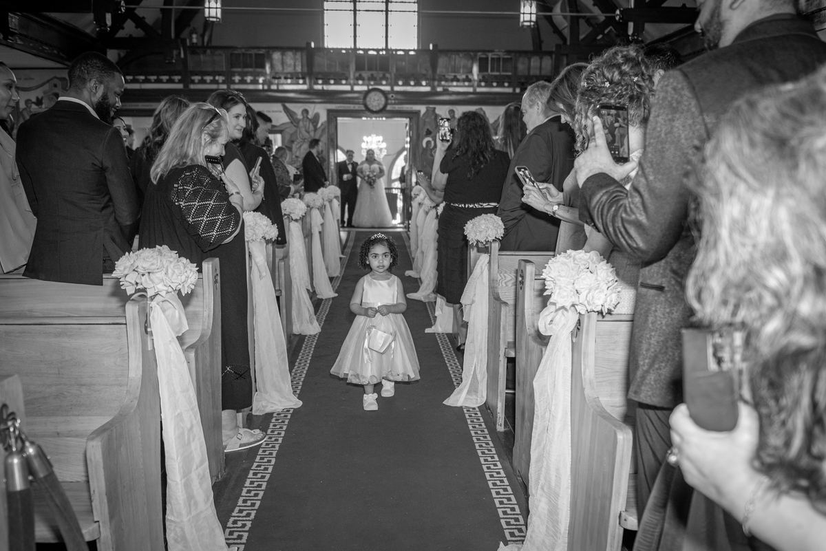 A young flower girl walks solemnly down the aisle in a dimly lit church with white floral decorations.