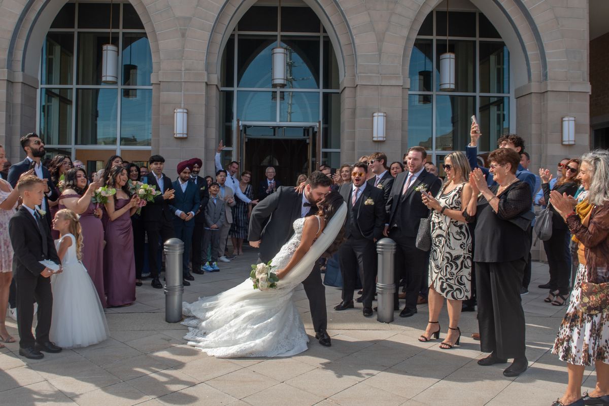 A groom lifts his bride in a dip outside a grand stone building as guests cheer and capture the moment with phones.