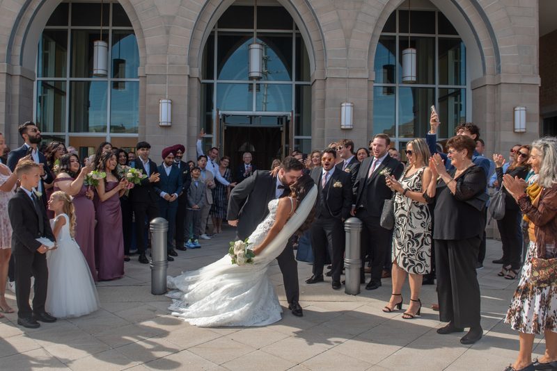 A groom lifts his bride in a dip outside a grand stone building as guests cheer and capture the moment with phones.