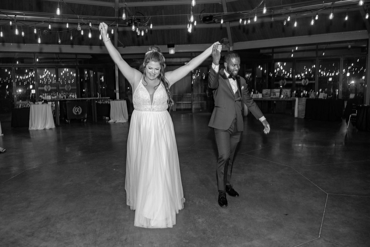 A bride and groom dance with both arms raised in a dim ballroom under strings of warm lights, captured in black and white.