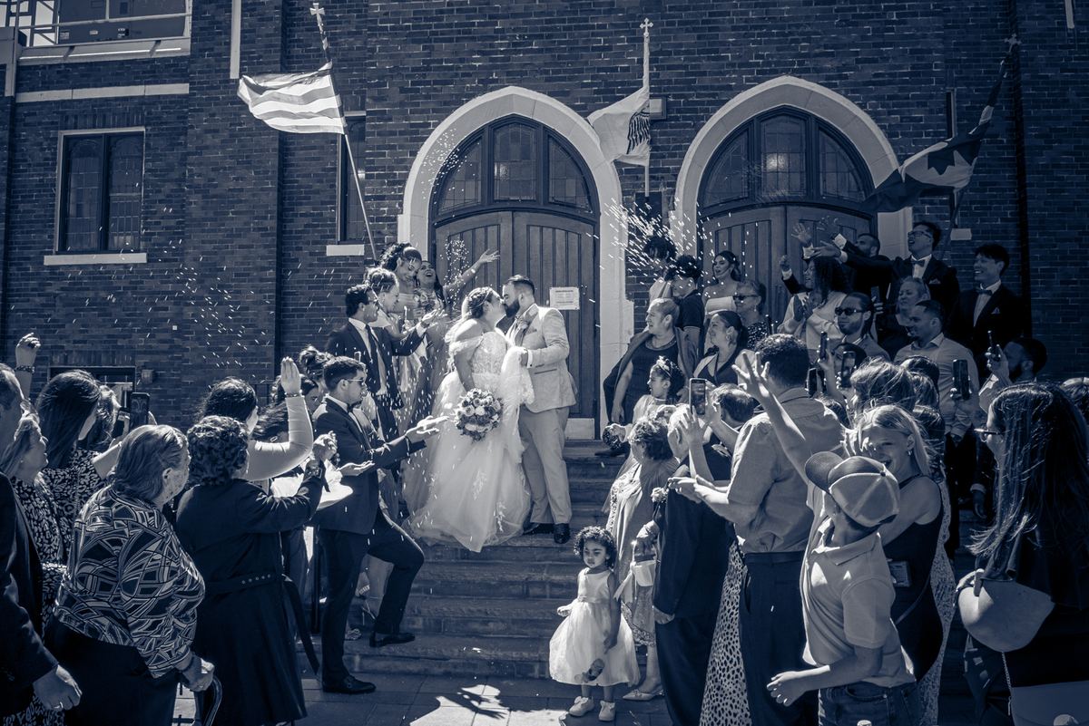 A newlywed couple descends steps, showered in confetti as guests cheer and raise phones, with a child in white nearby.