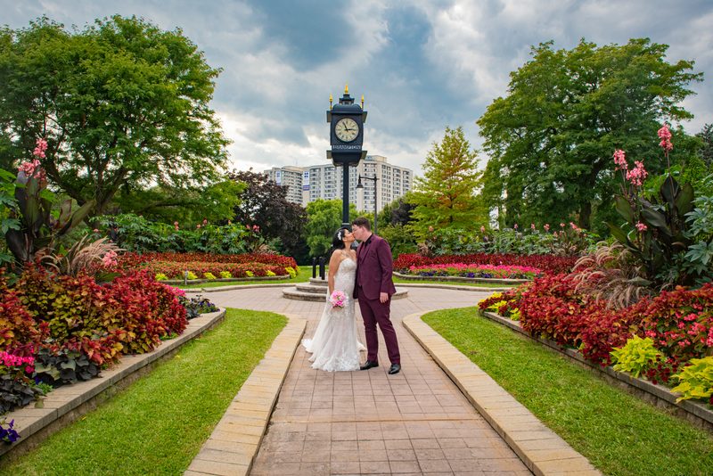 Bride and groom kiss along a garden path surrounded by flowers and greenery with a clock tower in the background.