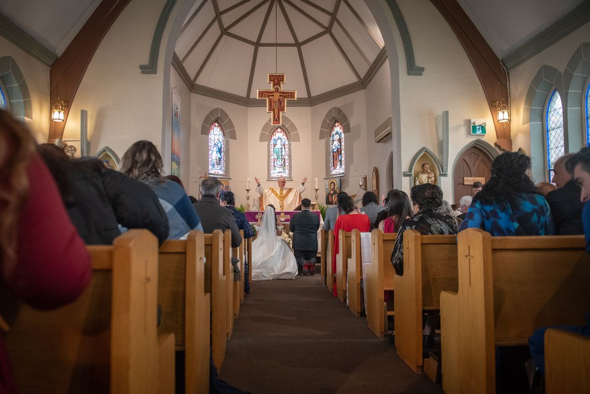 Bride and groom kneel at altar as priest blesses them in warmly lit church with stained-glass windows.