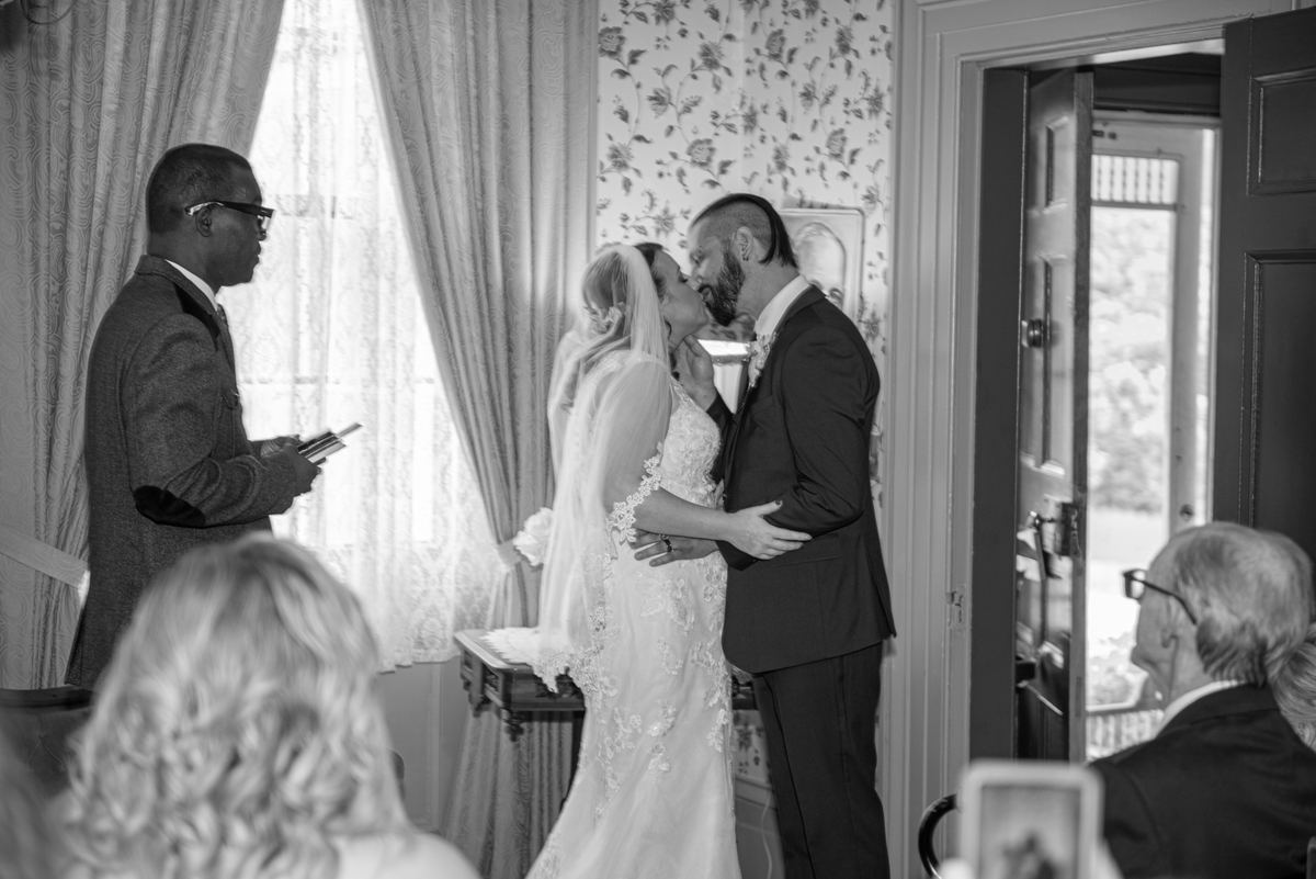 Bride and groom kiss during ceremony, framed by lace curtains and soft light, with guests observing.