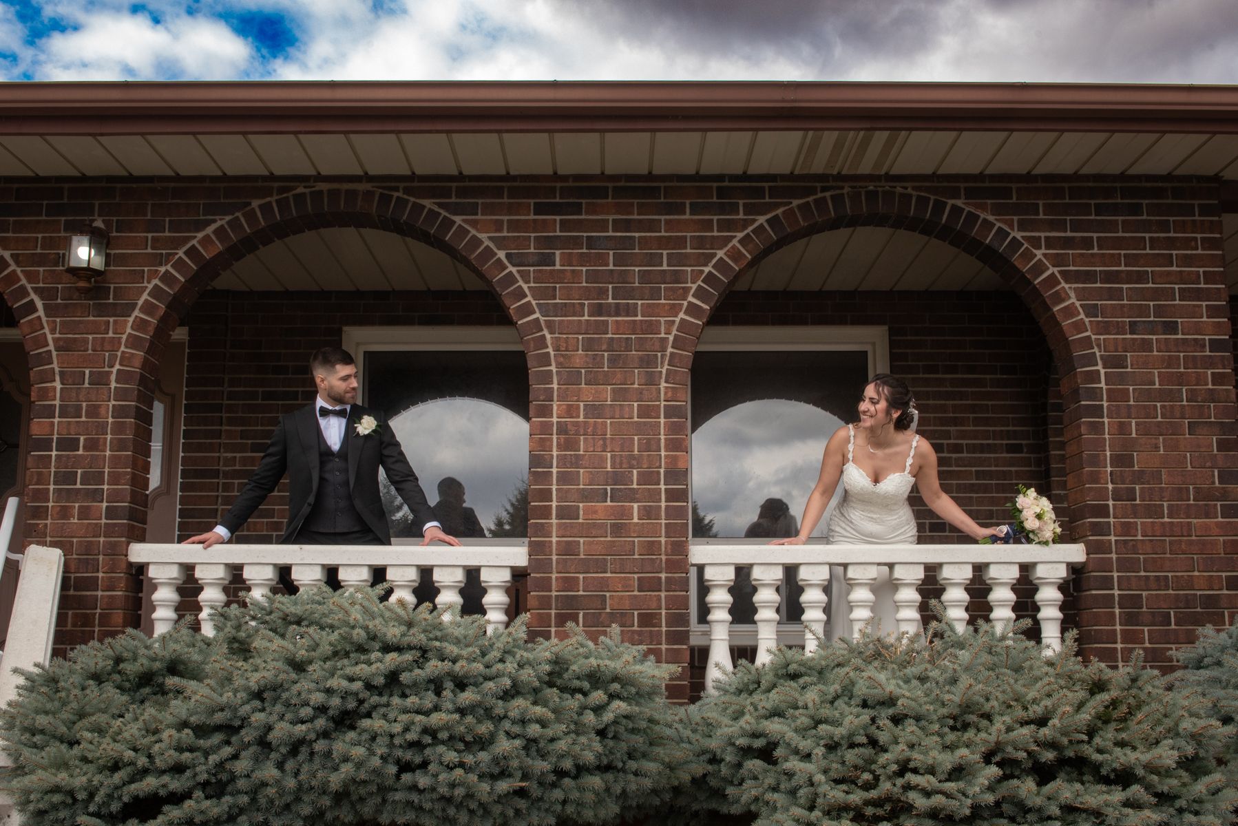 A smiling bride and groom stand on a brick balcony framed by arched windows, looking out at the camera.