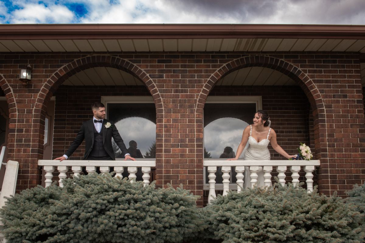 A smiling bride and groom stand on a brick balcony framed by arched windows, looking out at the camera.