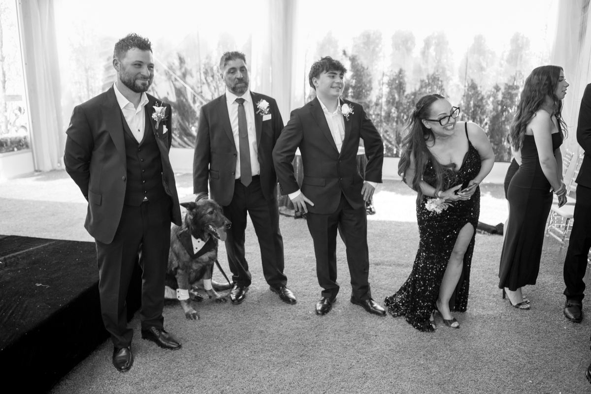 Groomsmen and a formally dressed dog stand beside a laughing woman in a sequined gown at a wedding reception.