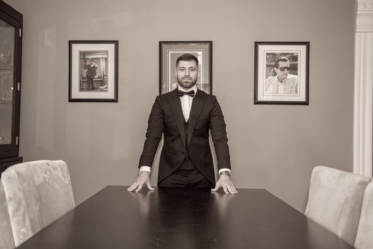 A formally dressed man in a tuxedo and bow tie stands behind a dark dining table at Danielle and Michael's wedding.