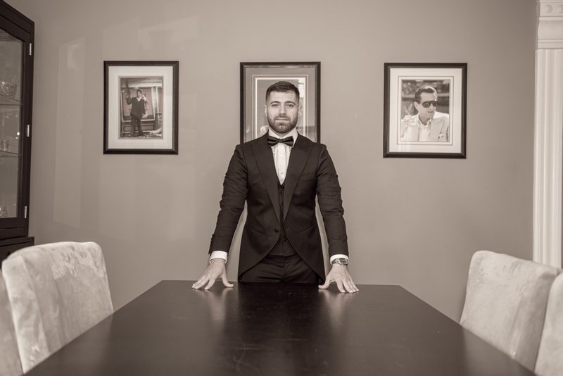 A formally dressed man in a tuxedo and bow tie stands behind a dark dining table at Danielle and Michael's wedding.