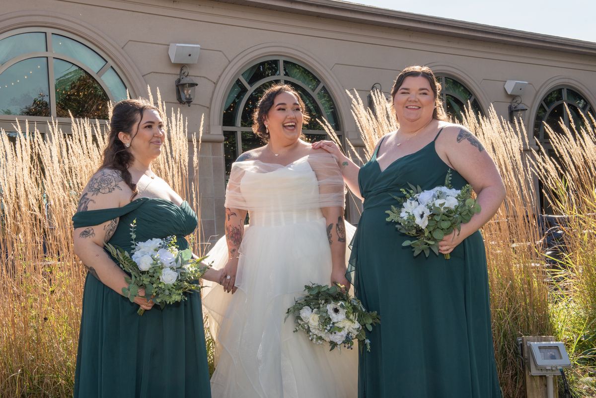 Four women, including a bride in white, stand outdoors smiling and laughing near ornamental grasses.