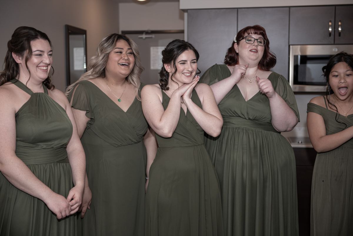 Five bridesmaids in olive-green dresses laugh joyfully in a neutral-toned kitchen with soft indoor lighting.