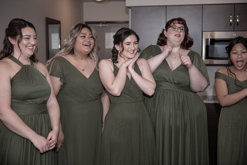 Five bridesmaids in olive-green dresses laugh joyfully in a neutral-toned kitchen with soft indoor lighting.