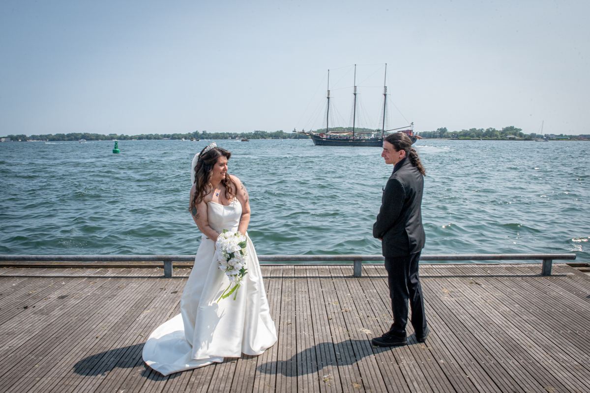 A bride in white and groom in a dark suit stand on a wooden dock by the water, smiling at each other.