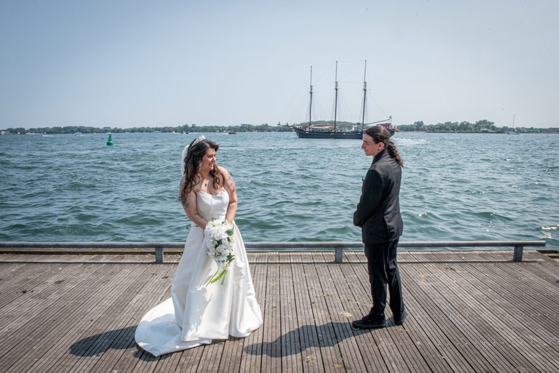 A bride in white and groom in a dark suit stand on a wooden dock by the water, smiling at each other.