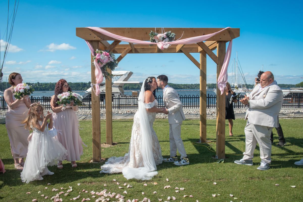 Bride and groom kiss under a pink-draped arbor at an outdoor ceremony with guests and waterfront view.