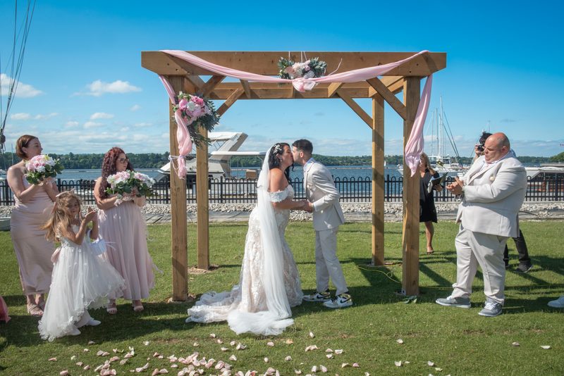 Bride and groom kiss under a pink-draped arbor at an outdoor ceremony with guests and waterfront view.