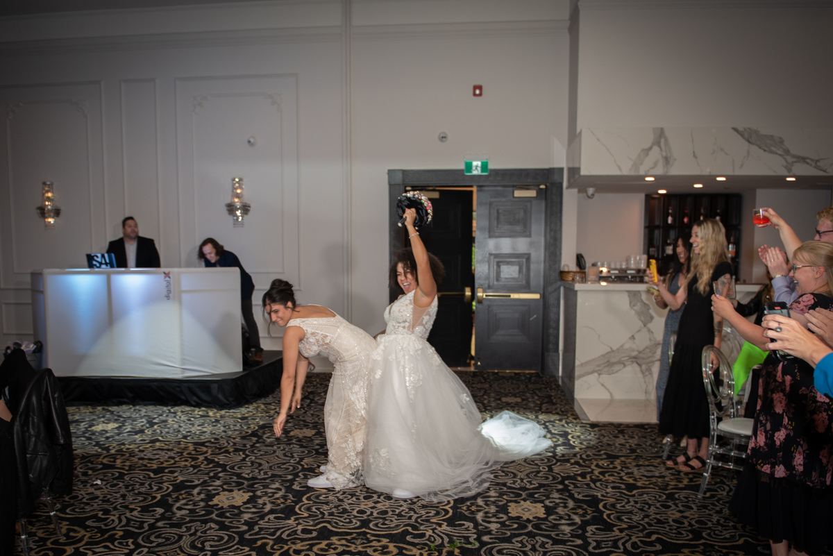 The bride tosses her bouquet as a bridesmaid attempts to catch it, with guests cheering in the background.