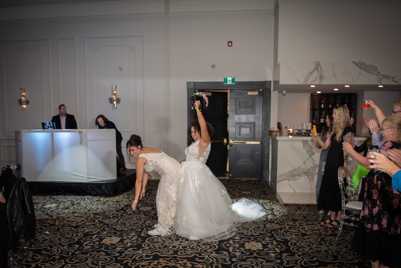 The bride tosses her bouquet as a bridesmaid attempts to catch it, with guests cheering in the background.