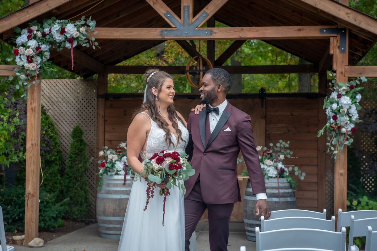 Alexandria and Derado stand beneath a rustic wooden arbor decorated with red and white flowers, smiling at each other.