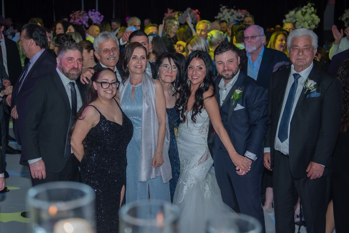 Bride and groom surrounded by smiling guests in formal wear, illuminated by event lighting and candles.