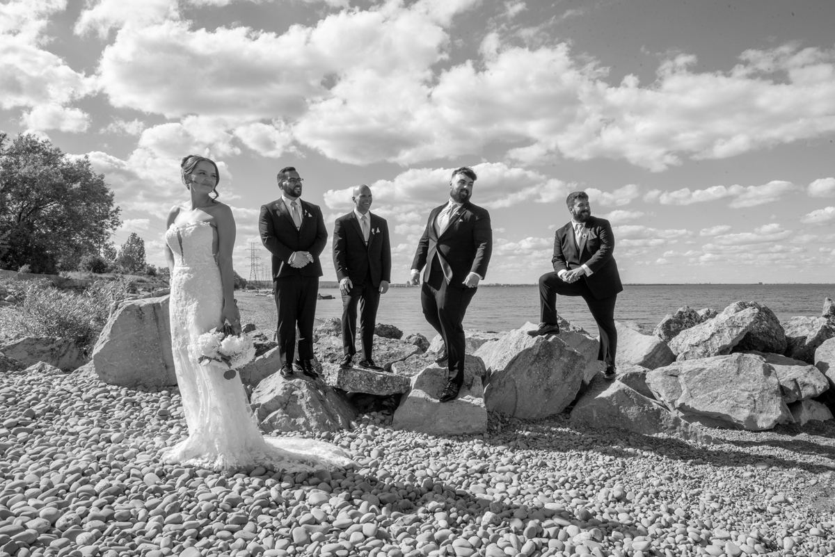A bride in a white gown stands with four groomsmen on a rocky shore under a bright sky, emphasizing natural light.