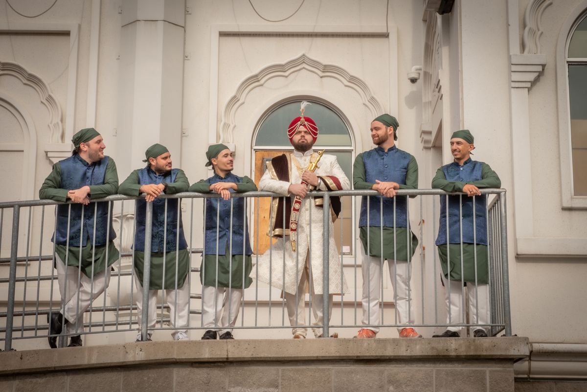A groom in white and red stands with four groomsmen in green and blue on a balcony overlooking a white building.