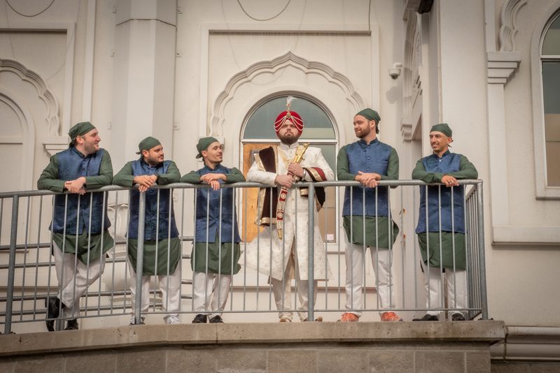 A groom in white and red stands with four groomsmen in green and blue on a balcony overlooking a white building.