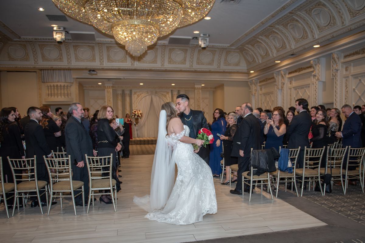 Cassandra and Brandon share a tender kiss at the end of their aisle, surrounded by guests in an opulent ballroom.