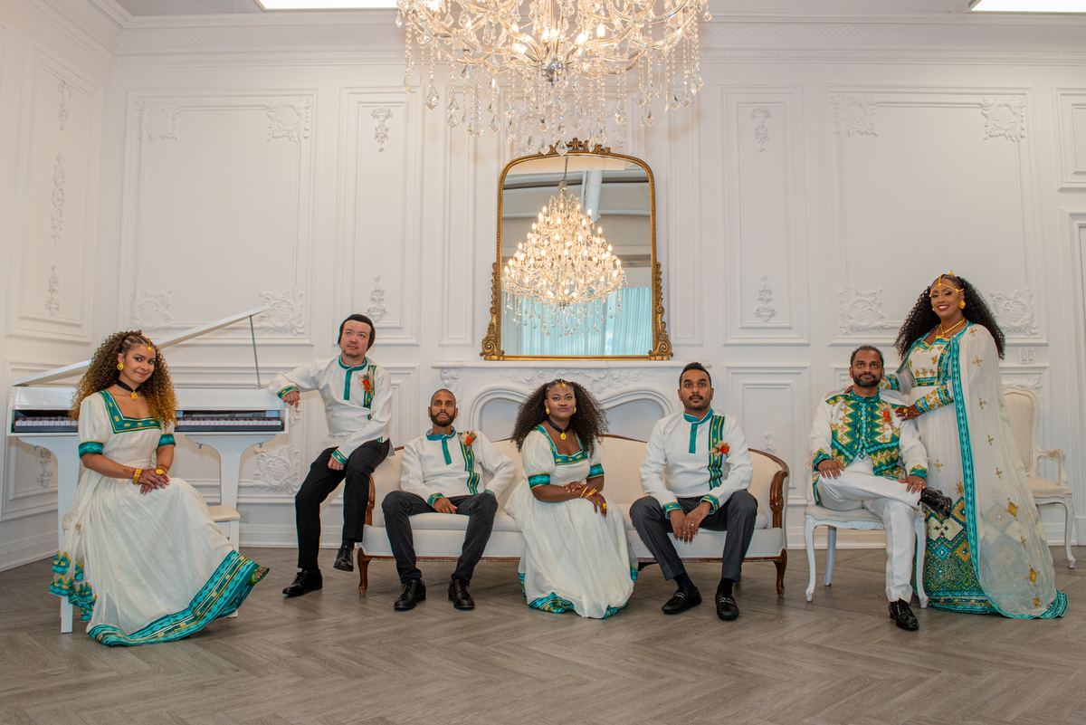 Seven people in white and teal performative attire pose formally in an ornate room with a chandelier.