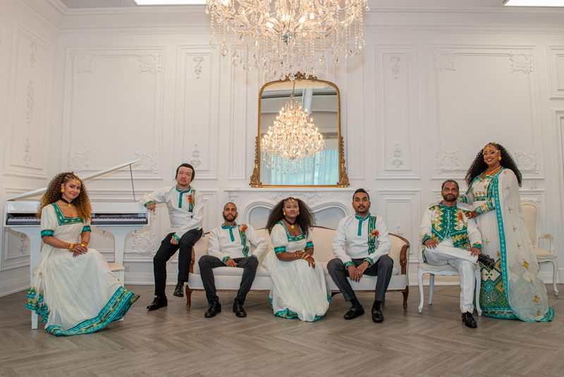 Seven people in white and teal performative attire pose formally in an ornate room with a chandelier.