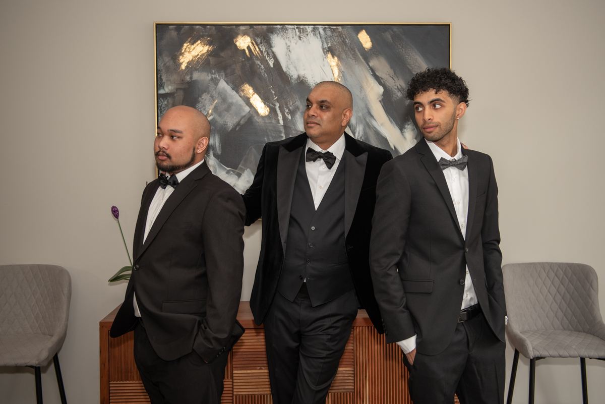 Three formally dressed men stand together in a modern room with soft lighting and an abstract painting behind them.