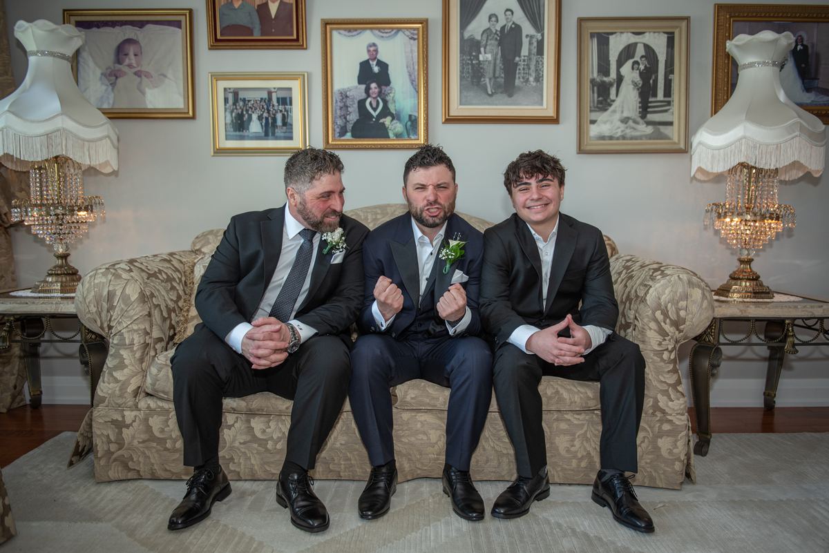 Three men in suits sit on a patterned sofa, laughing together in an elegantly decorated room with framed photos.