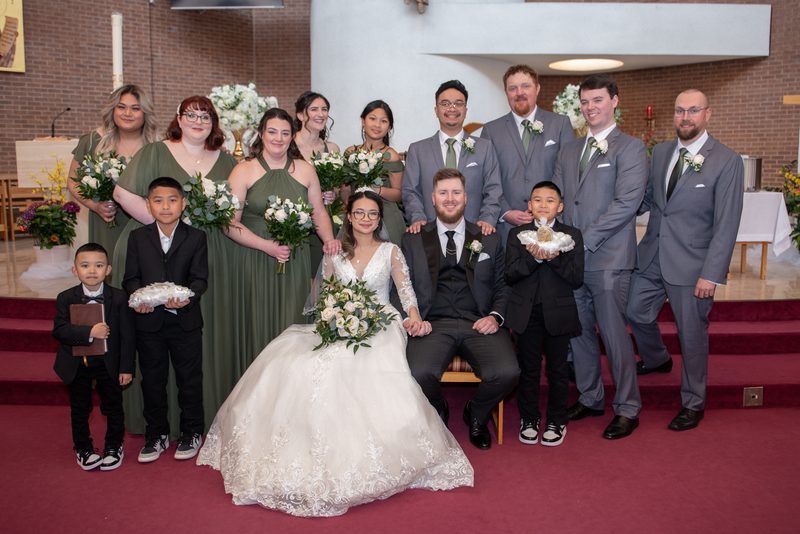 Bride and groom sit center with bridesmaids in olive green, groomsmen in gray suits, and two ring bearers holding baskets.