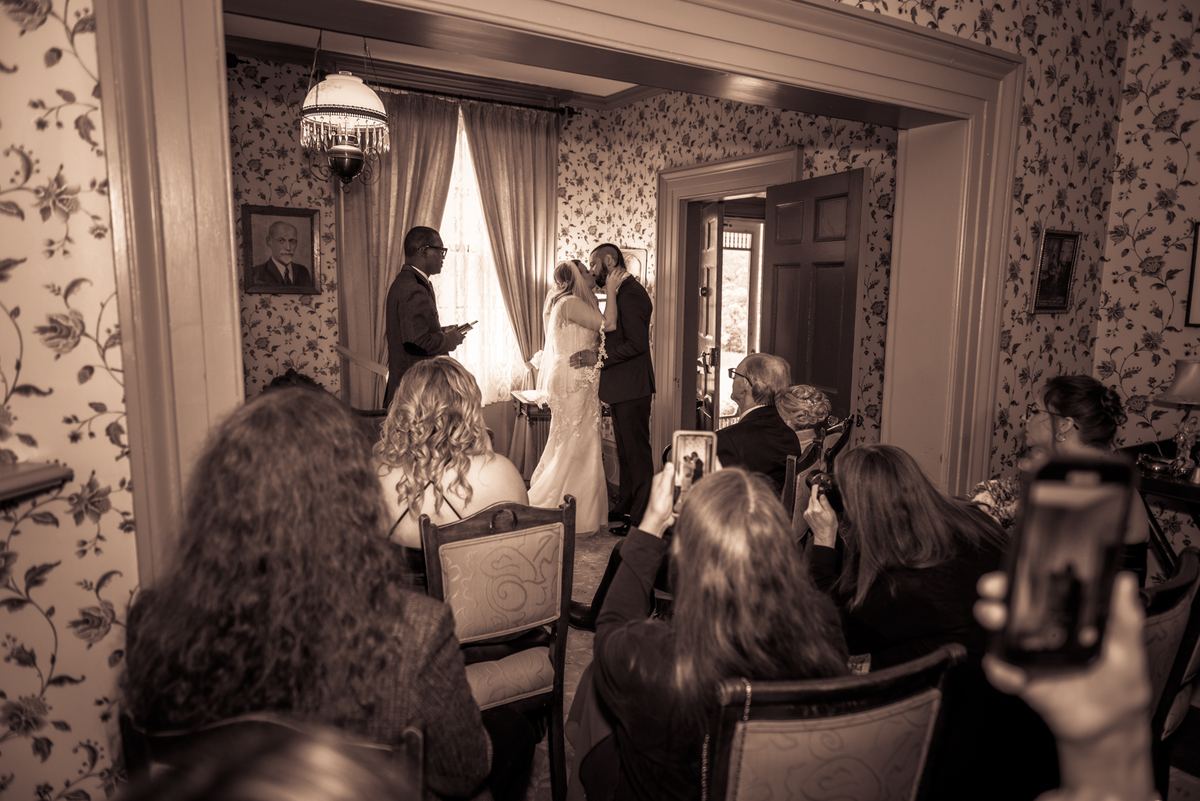 Bride and groom kiss in vintage room with floral wallpaper and natural light, guests watching on phones.