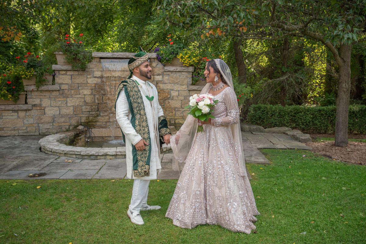 A smiling South Asian bride and groom in traditional attire stand hand-in-hand by a stone fountain surrounded by greenery.