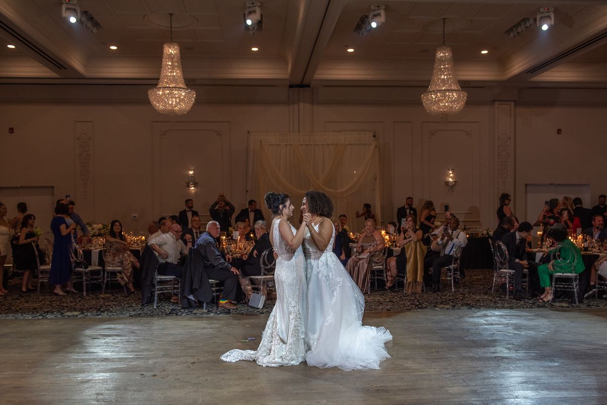 Two brides dance closely in a warmly lit ballroom surrounded by candlelit tables and guests, celebrating their union.