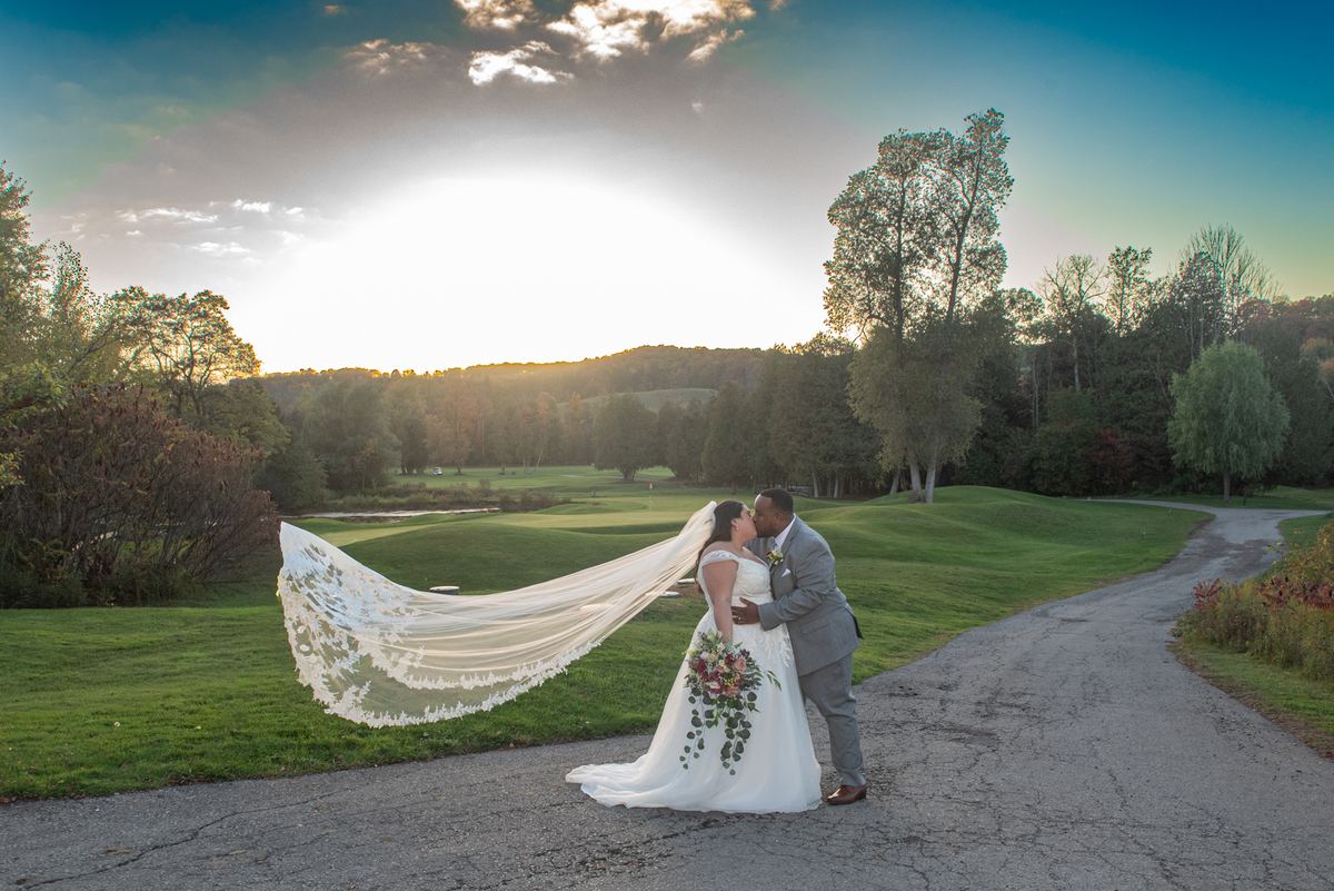 Bride and groom kiss beside a golf course path as the sun sets, her lace veil billowing in the wind.