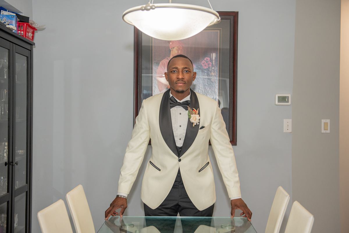 A man in a cream tuxedo stands behind a glass dining table in a formal setting, hands resting on the edge.