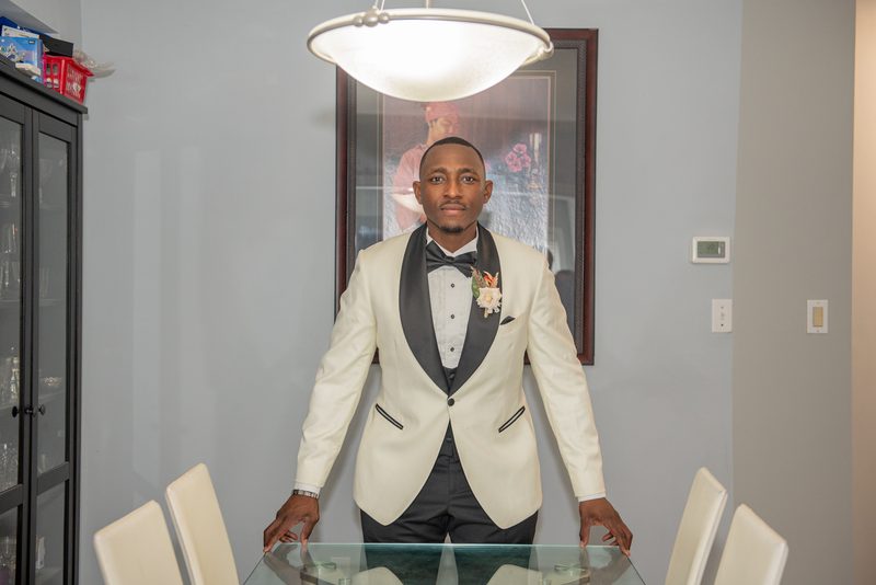 A man in a cream tuxedo stands behind a glass dining table in a formal setting, hands resting on the edge.