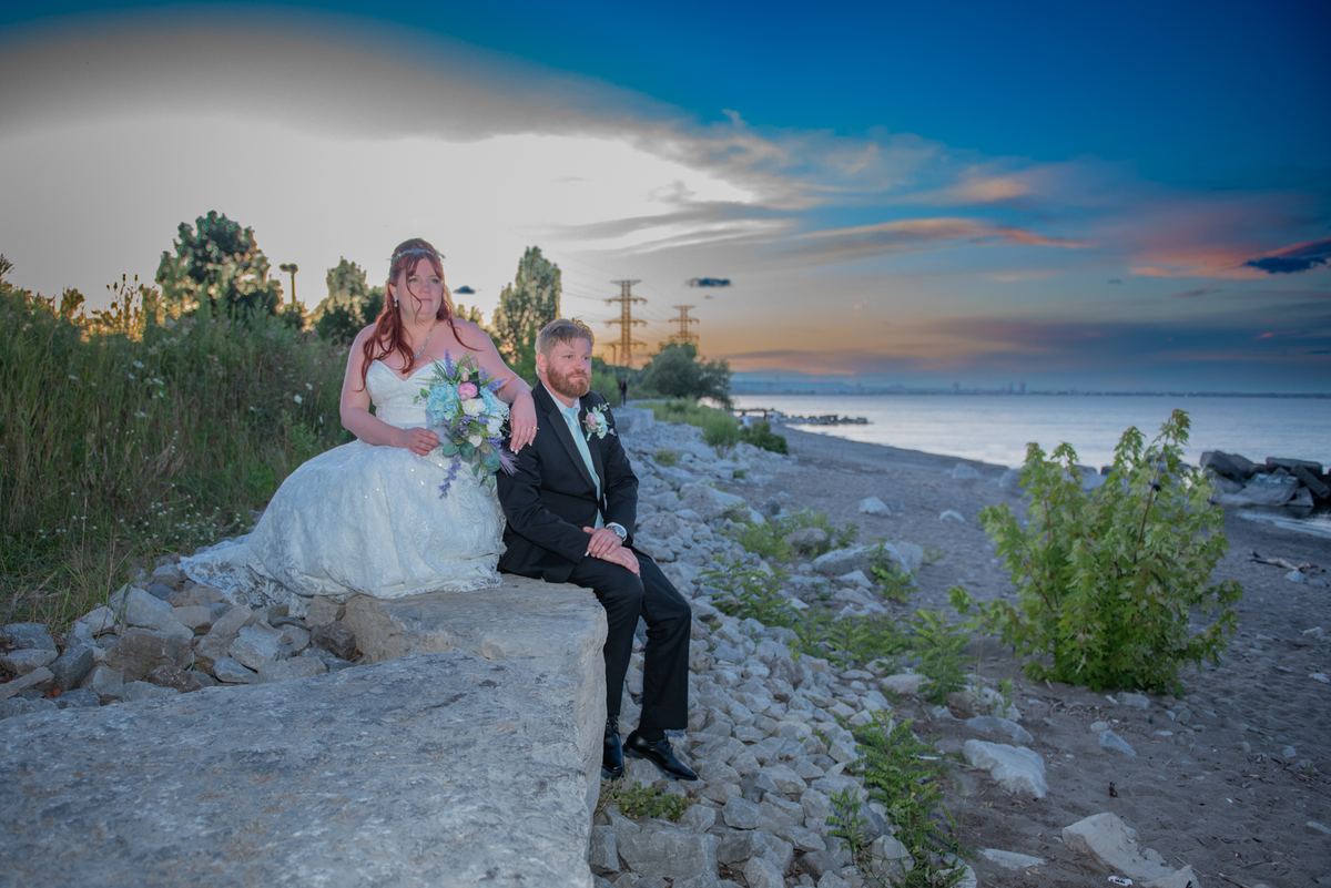 A bride and groom sit on a stone embankment by the waterfront at dusk, holding hands with the city skyline in the background.