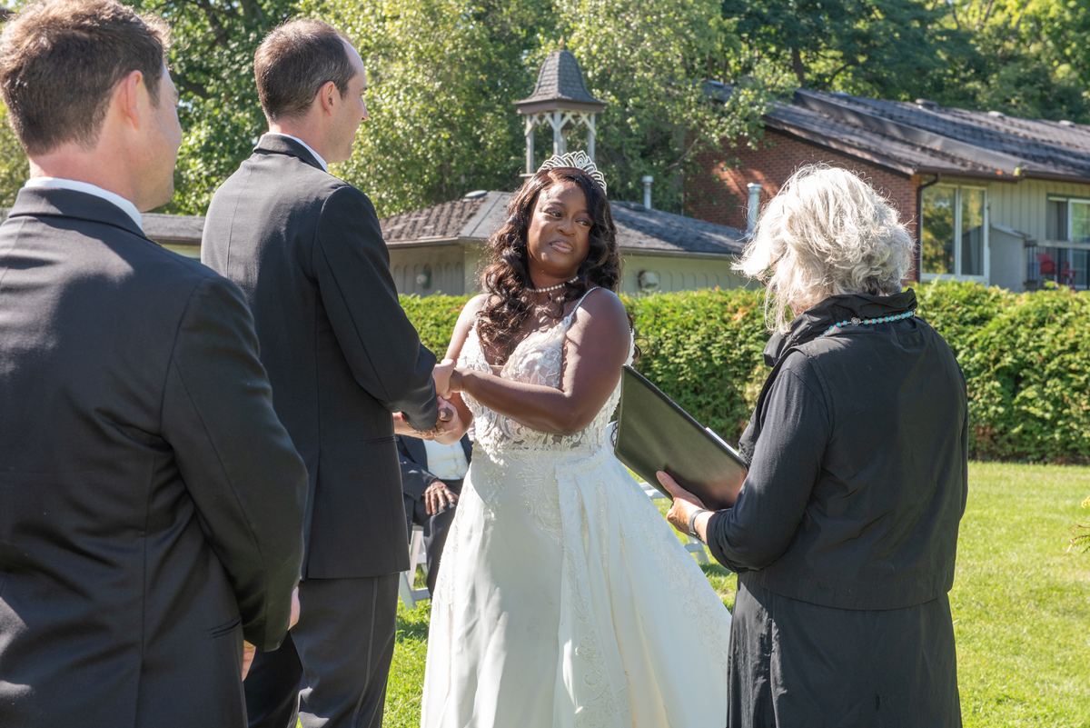 A smiling bride in a tiara exchanges rings with her groom under bright sunlight, surrounded by greenery.