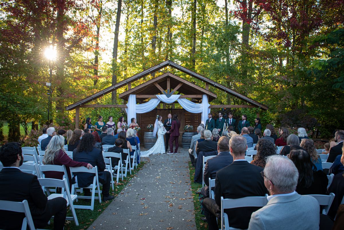 Bride and groom stand at an altar in a sunlit woodland clearing, surrounded by autumn leaves and guests.