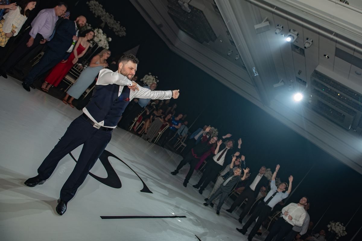 A formally dressed man performs an energetic dance move on a white floor, guests cheering around him under bright lights.