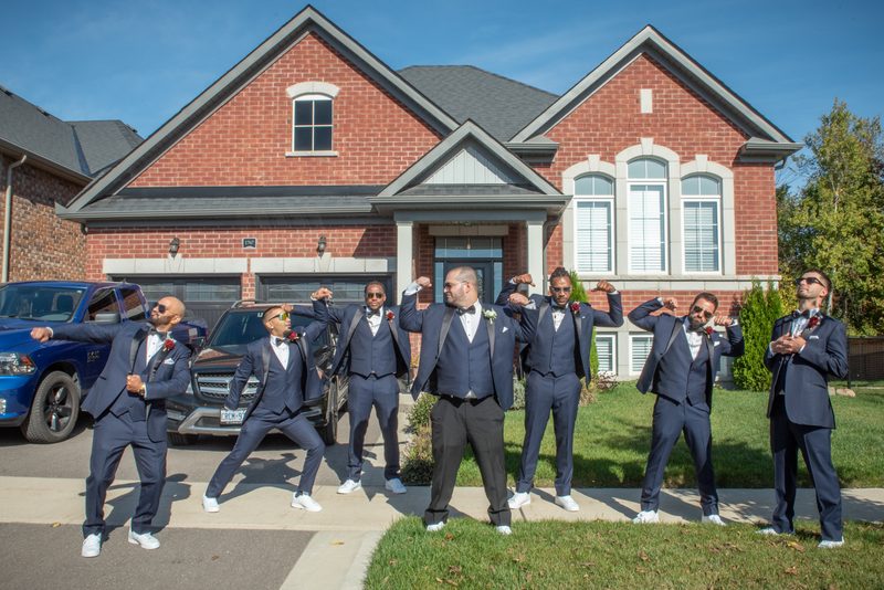 A groom and his groomsmen pose in synchronized, playful stances in front of a red-brick house on a sunny afternoon.