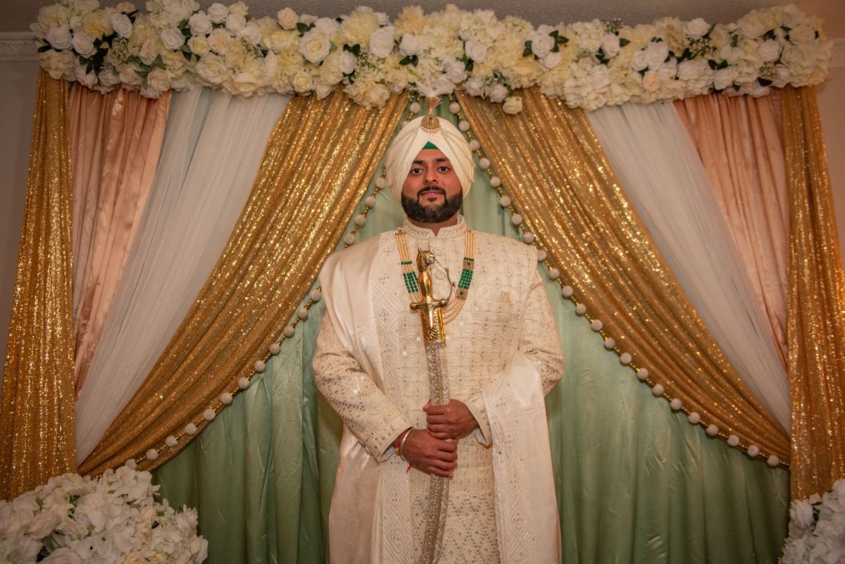 A formally dressed groom in white turban and embroidered sherwani holds a ceremonial sword against gold and white drapes.